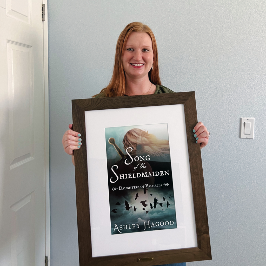 Ashley holding a framed print of her first book Song of the Shieldmaiden